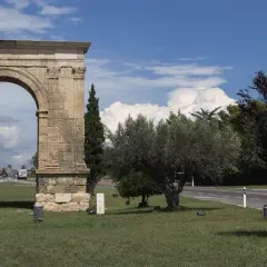 Arco romano de piedra, parte de una antigua roda de bara, flanqueado por árboles y palmeras bajo un cielo azul con nubes.