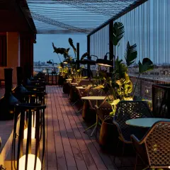 Terraza del Novotel Valencia con mesas y sillas de mimbre, iluminada por lámparas y rodeada de vegetación.