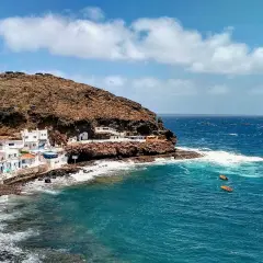 Pueblo costero blanco anidado en la ladera rocosa de **Tufia**. El mar azul turquesa choca contra las rocas, con pequeñas embarcaciones flotando.