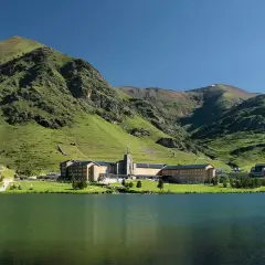 Lago sereno frente a un complejo de edificios en Queralbs, rodeado de montañas verdes y un cielo azul claro.