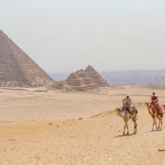 Un viaje a Egipto inolvidable: personas montan camellos frente a las pirámides de Giza bajo un cielo brumoso.