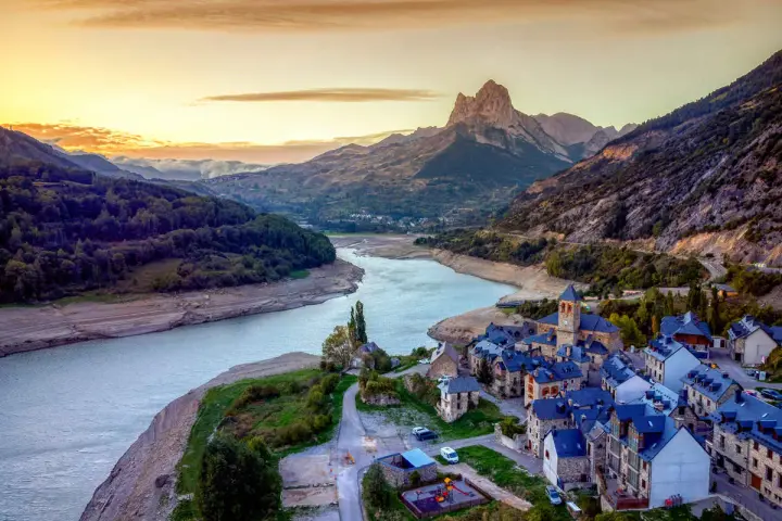 Un pueblo de piedra con tejados azules a orillas de un embalse, bajo un pico montañoso imponente. Un ejemplo de los pueblos mas bonitos de Huesca.