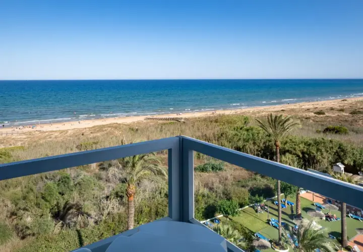 Vista desde balcón de hotel, mar azul, playa con gente y vegetación. Opiniones de hotel Playas de Guardamar positivas.