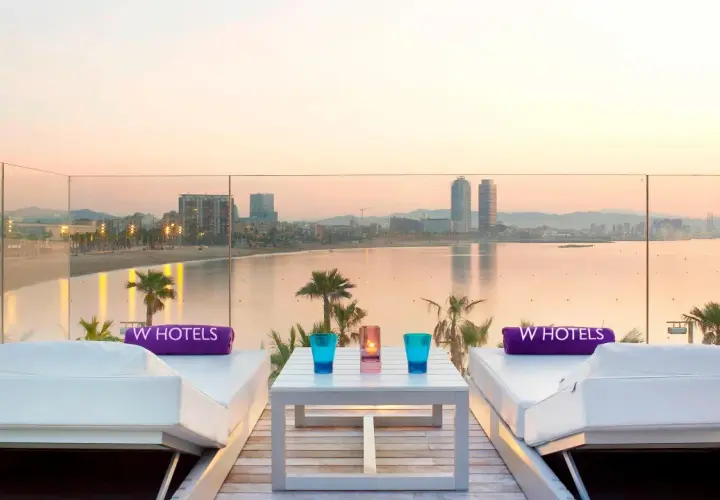Dos tumbonas blancas con toallas moradas de W Hotels, una mesa con vasos y velas, y vistas al mar y la ciudad de Barcelona al atardecer.
