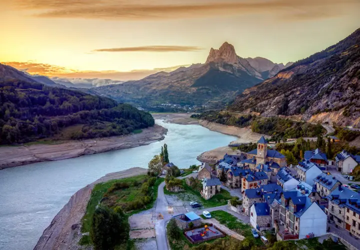 Un pueblo de piedra con tejados azules a orillas de un embalse, bajo un pico montañoso imponente. Un ejemplo de los pueblos mas bonitos de Huesca.