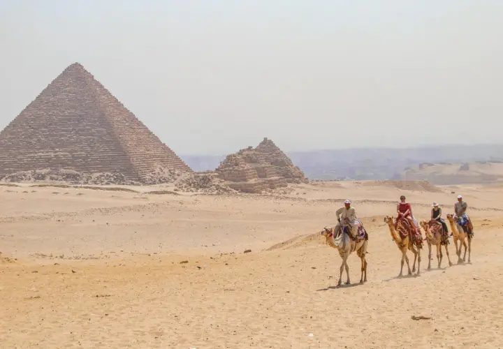 Un viaje a Egipto inolvidable: personas montan camellos frente a las pirámides de Giza bajo un cielo brumoso.