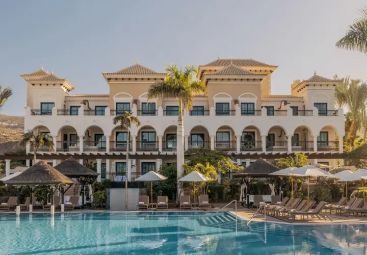 Piscina y edificio del Gran Meliá Palacio de Isora, con palmeras y tumbonas bajo el sol.