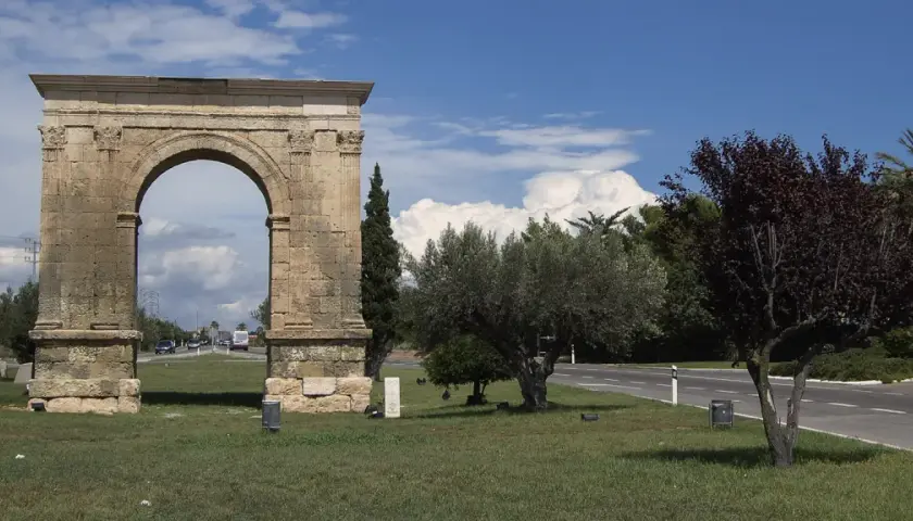 Arco romano de piedra, parte de una antigua roda de bara, flanqueado por árboles y palmeras bajo un cielo azul con nubes.