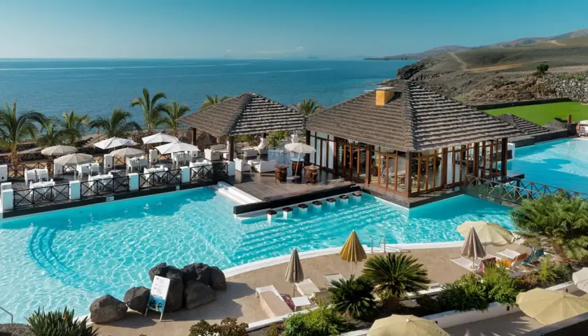 Un resort todo incluido con piscinas cristalinas, palmeras y vistas al mar. Mesas al aire libre y un bar acuático invitan a relajarse bajo el sol.