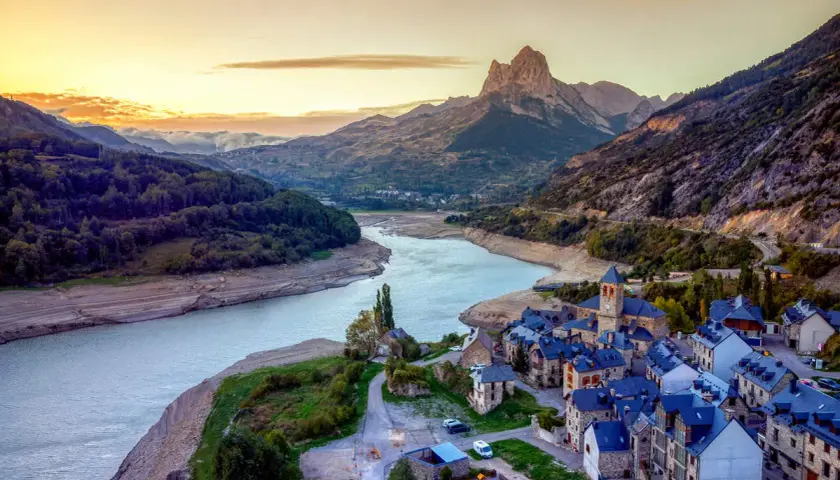 Un pueblo de piedra con tejados azules a orillas de un embalse, bajo un pico montañoso imponente. Un ejemplo de los pueblos mas bonitos de Huesca.
