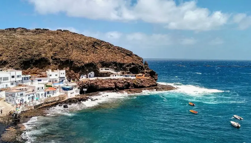 Pueblo costero blanco anidado en la ladera rocosa de **Tufia**. El mar azul turquesa choca contra las rocas, con pequeñas embarcaciones flotando.