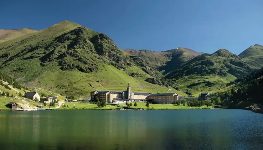 Lago sereno frente a un complejo de edificios en Queralbs, rodeado de montañas verdes y un cielo azul claro.