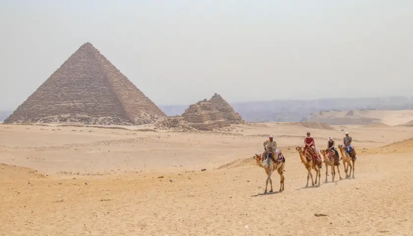 Un viaje a Egipto inolvidable: personas montan camellos frente a las pirámides de Giza bajo un cielo brumoso.