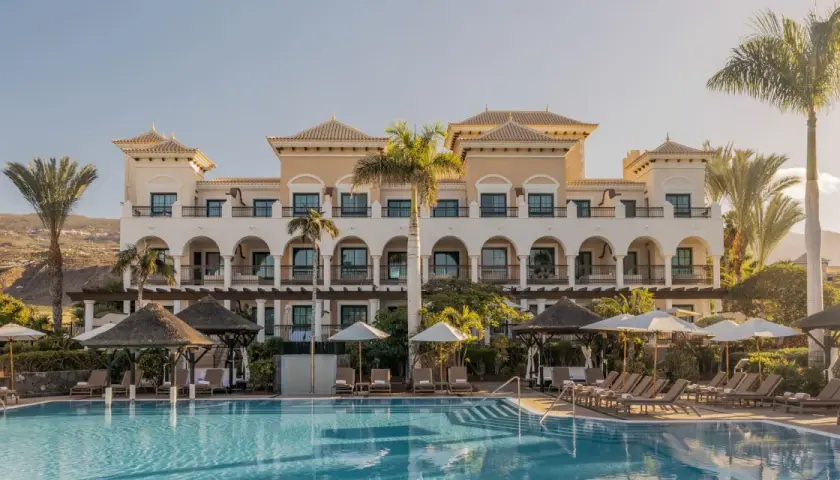 Piscina y edificio del Gran Meliá Palacio de Isora, con palmeras y tumbonas bajo el sol.
