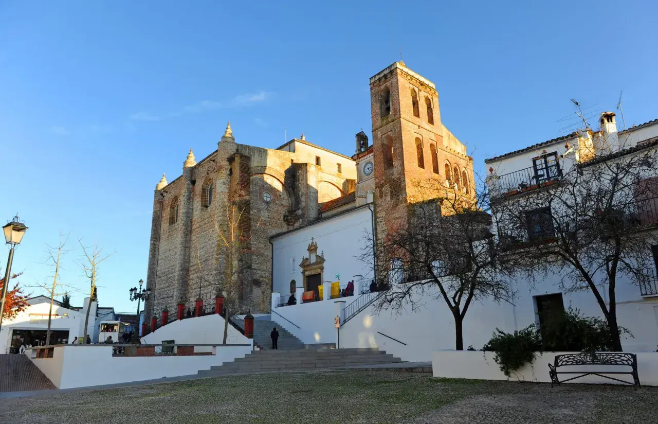 Iglesia de la Asunción en Cazalla de la Sierra, con su torre campanario y fachadas de piedra y cal bajo un cielo azul.