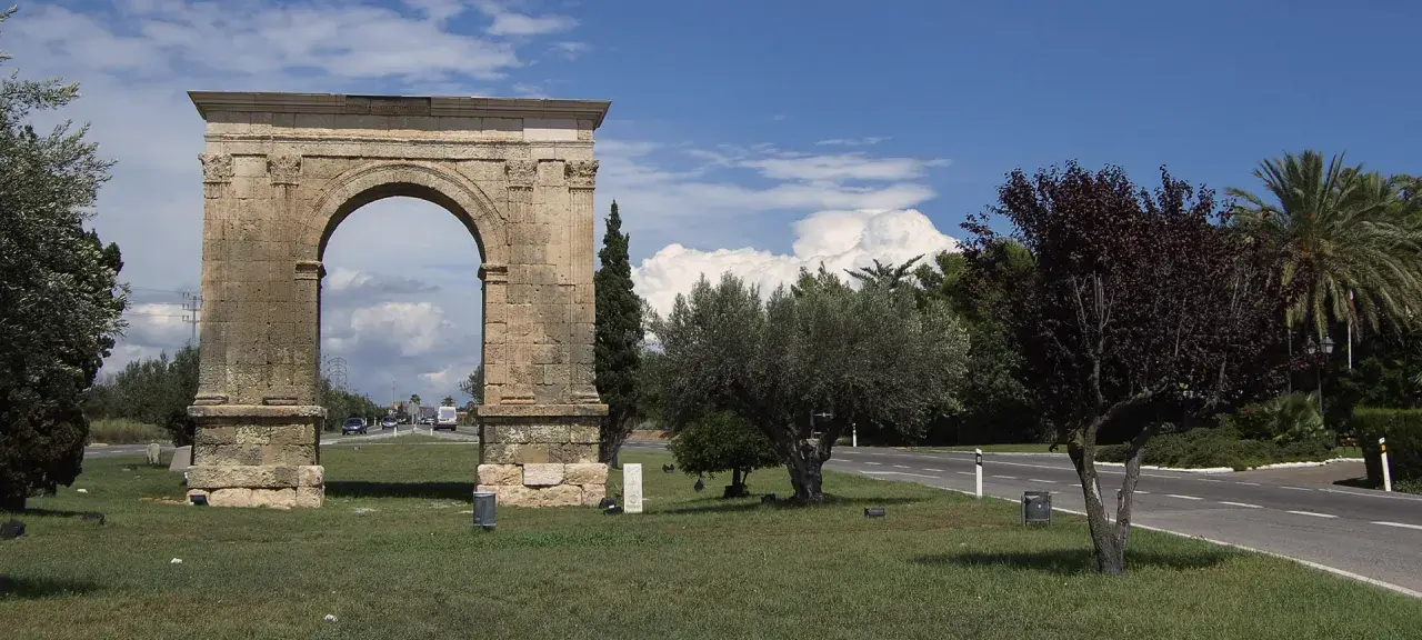 Arco romano de piedra, parte de una antigua roda de bara, flanqueado por árboles y palmeras bajo un cielo azul con nubes.