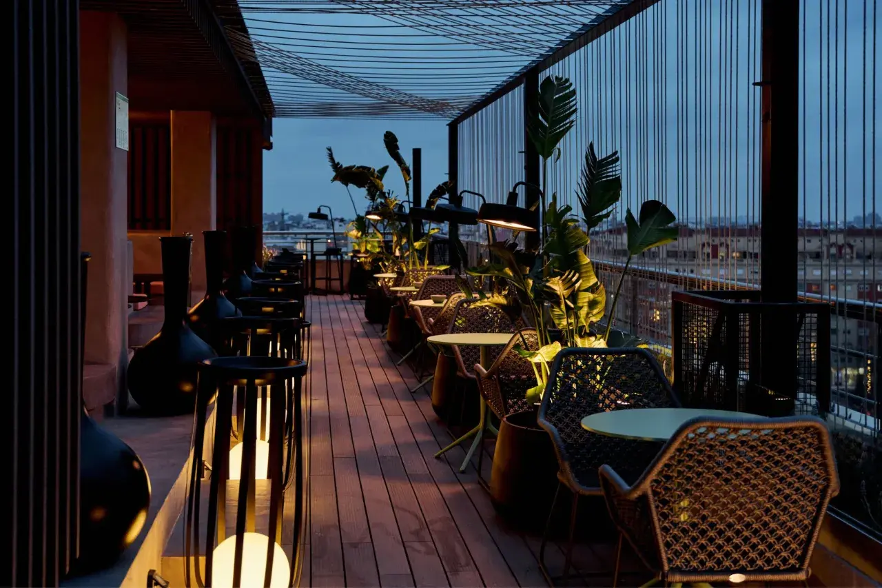Terraza del Novotel Valencia con mesas y sillas de mimbre, iluminada por lámparas y rodeada de vegetación.