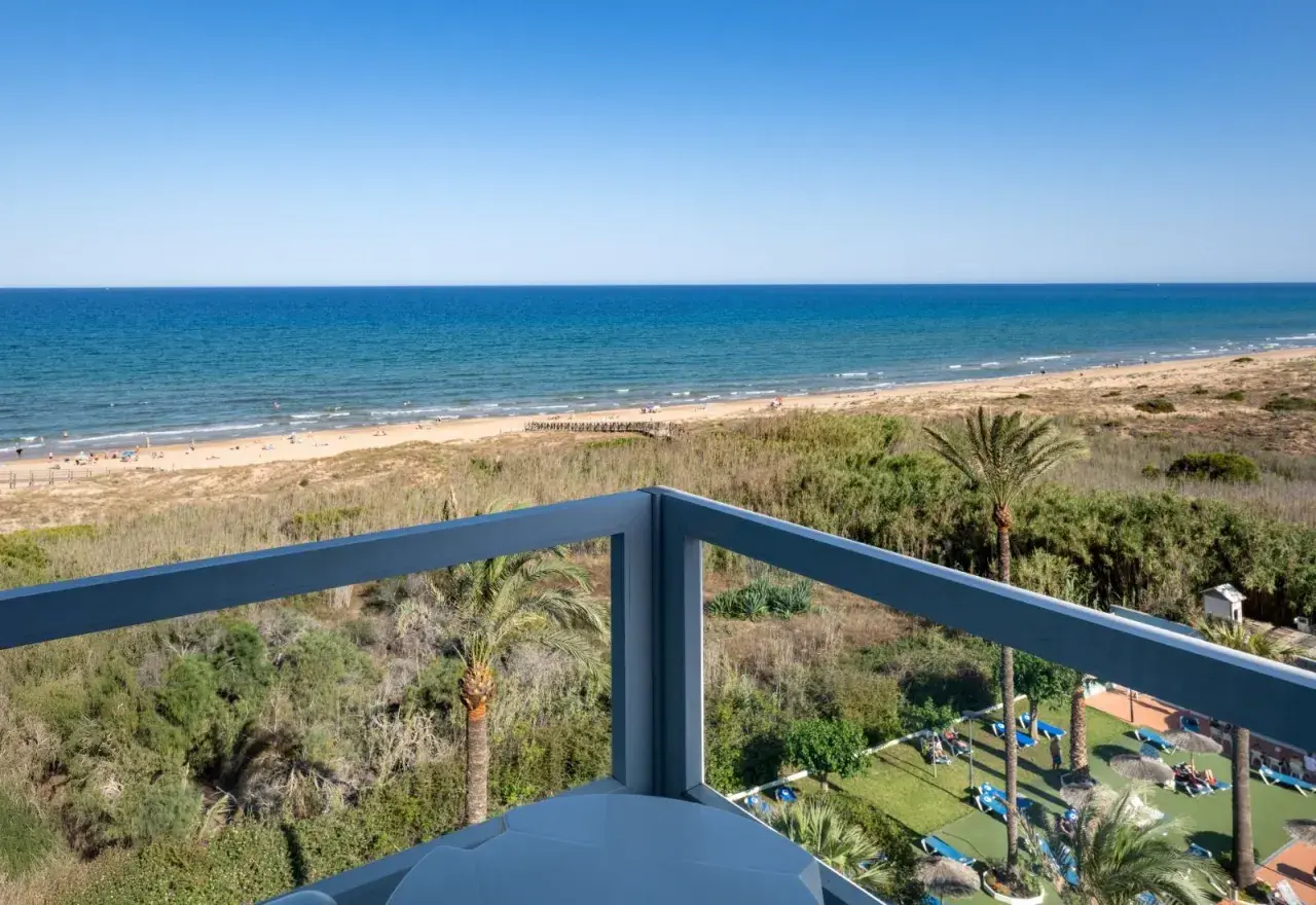 Vista desde balcón de hotel, mar azul, playa con gente y vegetación. Opiniones de hotel Playas de Guardamar positivas.