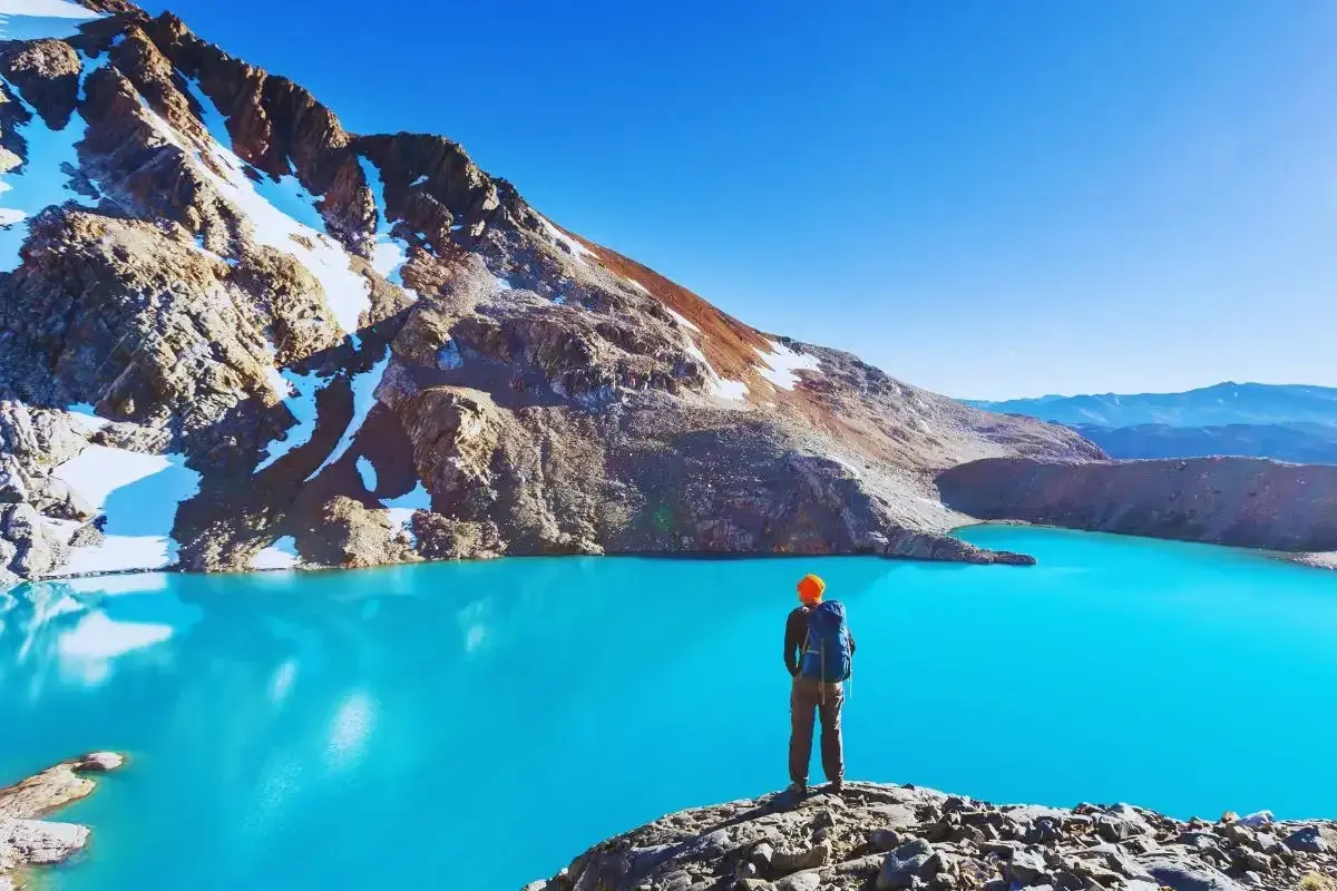 Sender solitario contempla un lago turquesa en la patagonia chilena, rodeado de montañas nevadas y un cielo azul intenso.