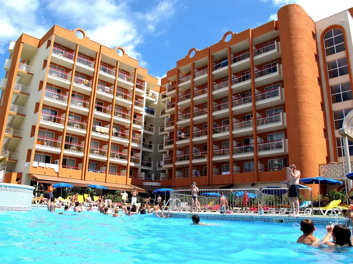 Gente disfrutando en la piscina del hotel Belvedere Salou, con edificios naranjas y blancos de fondo.