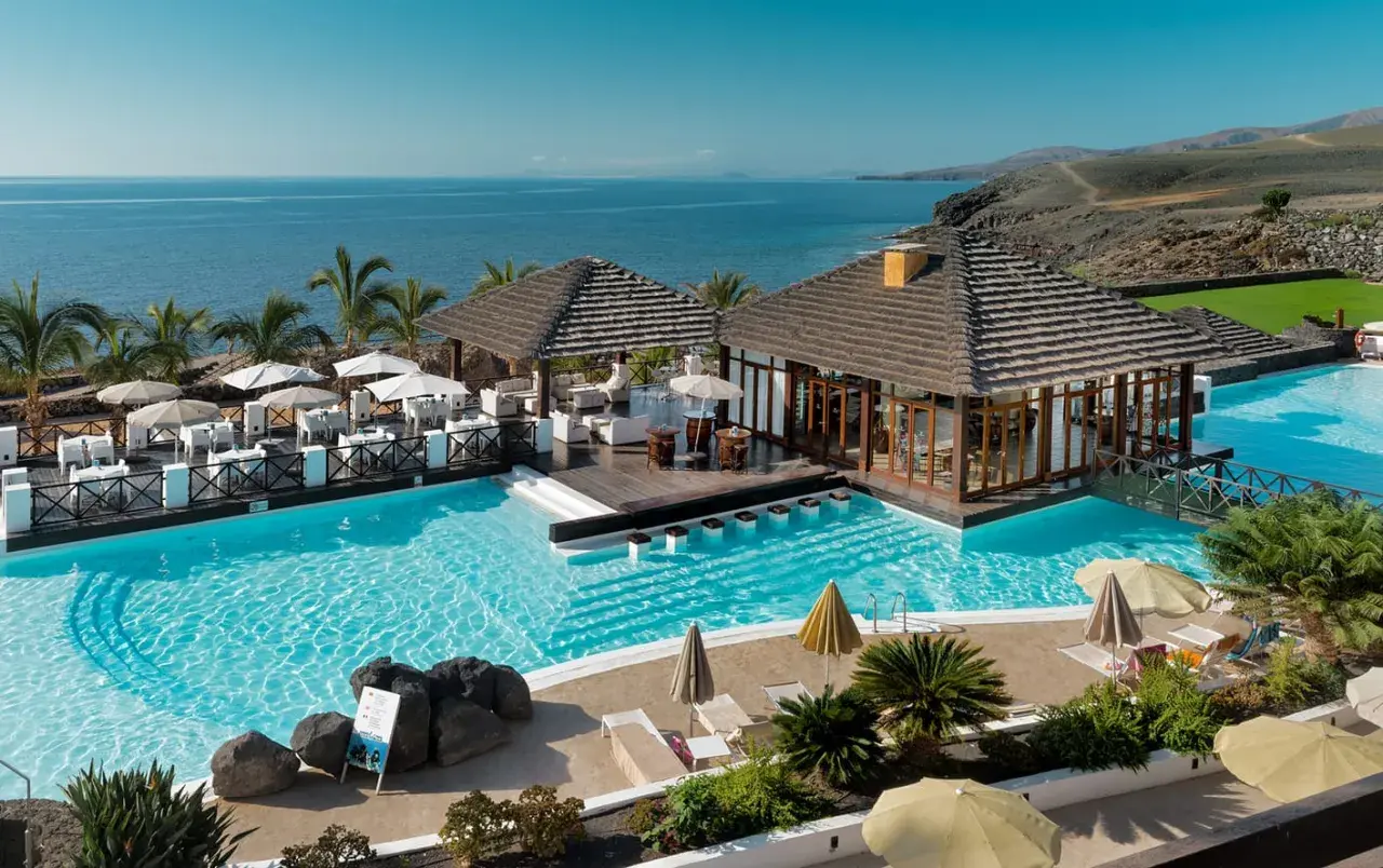 Un resort todo incluido con piscinas cristalinas, palmeras y vistas al mar. Mesas al aire libre y un bar acuático invitan a relajarse bajo el sol.