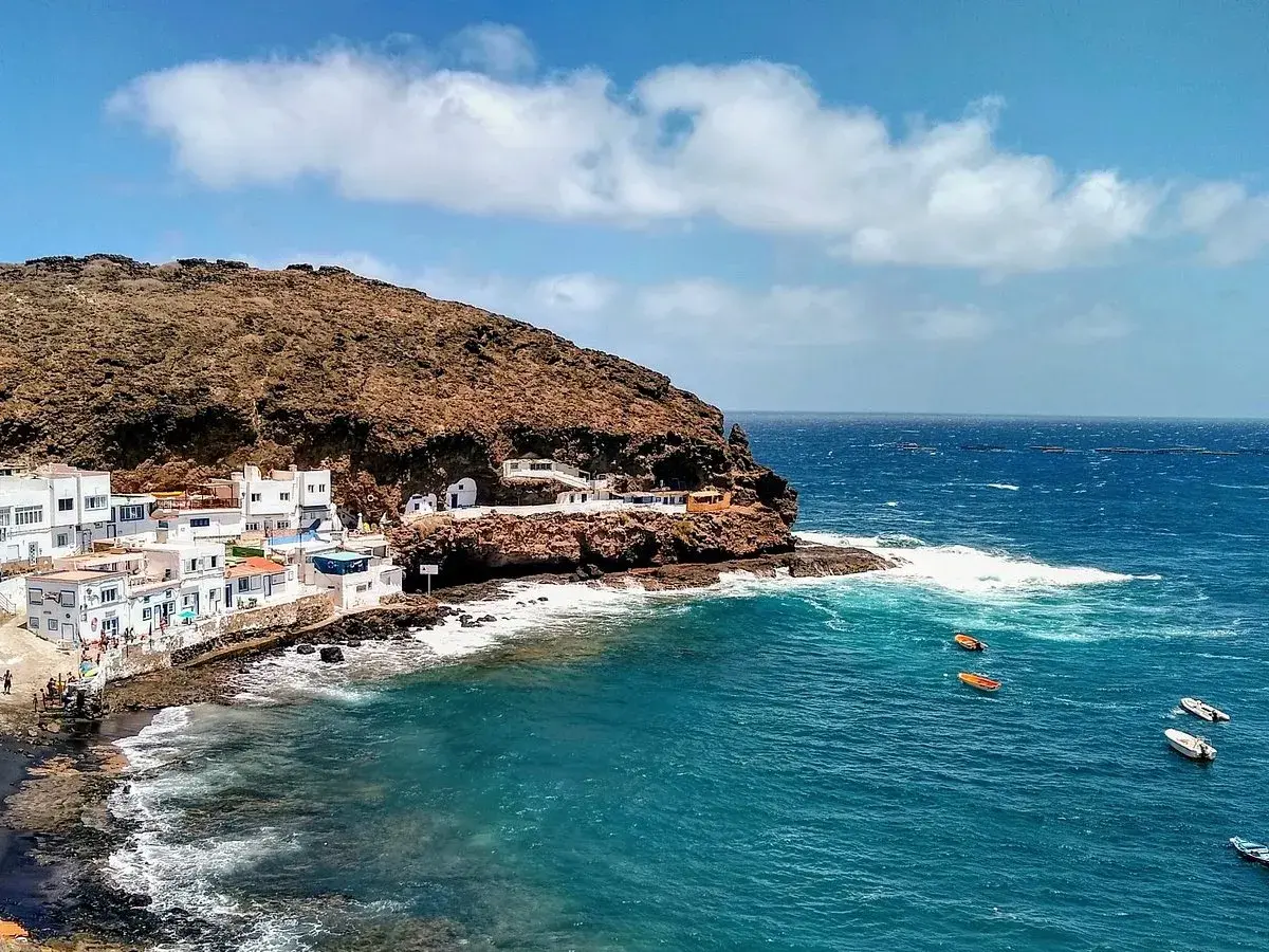 Pueblo costero blanco anidado en la ladera rocosa de **Tufia**. El mar azul turquesa choca contra las rocas, con pequeñas embarcaciones flotando.