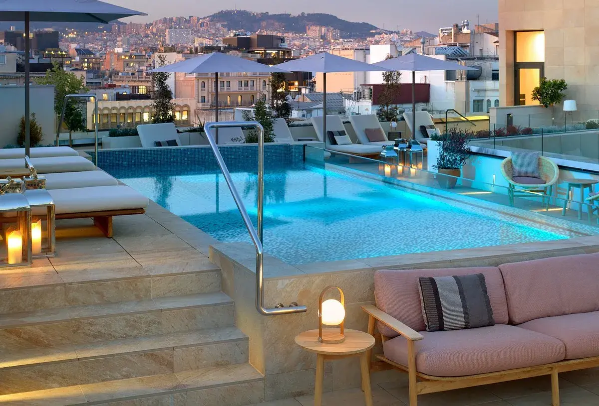 Piscina en la azotea del Barceló Raval con vistas a la ciudad al atardecer. Tumbonas, sofás y luces crean un ambiente acogedor.
