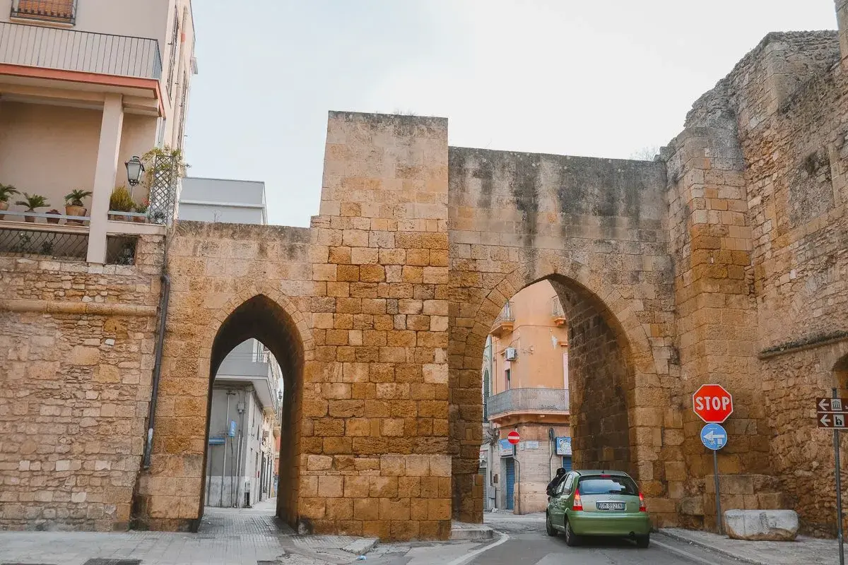 Antigua puerta de piedra con arcos, un coche verde pasa. Un lugar perfecto para un brindis por la historia.