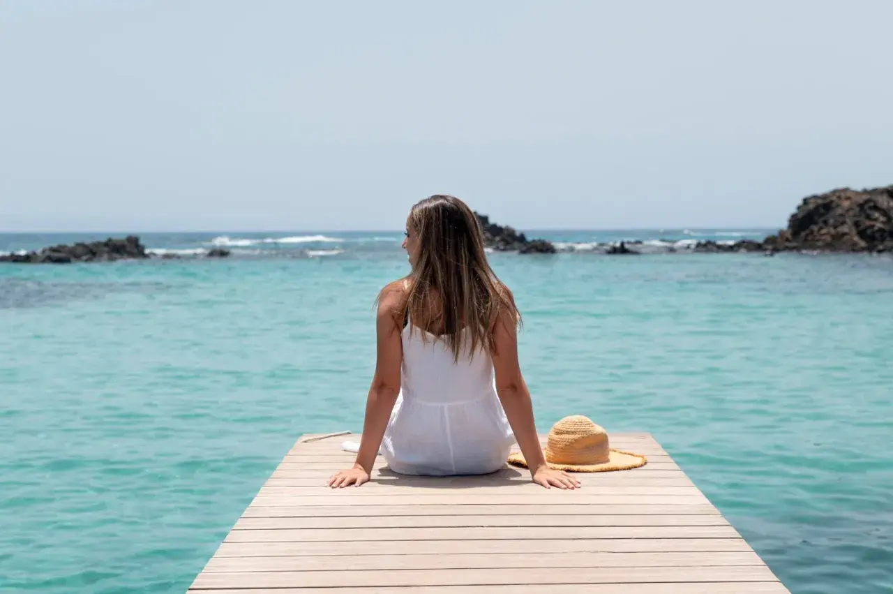 Mujer con vestido blanco sentada en un muelle de madera, disfrutando de la vista al mar turquesa. Su sombrero de paja descansa a su lado, perfecta para viajar sola.