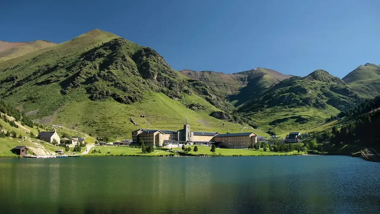 Lago sereno frente a un complejo de edificios en Queralbs, rodeado de montañas verdes y un cielo azul claro.
