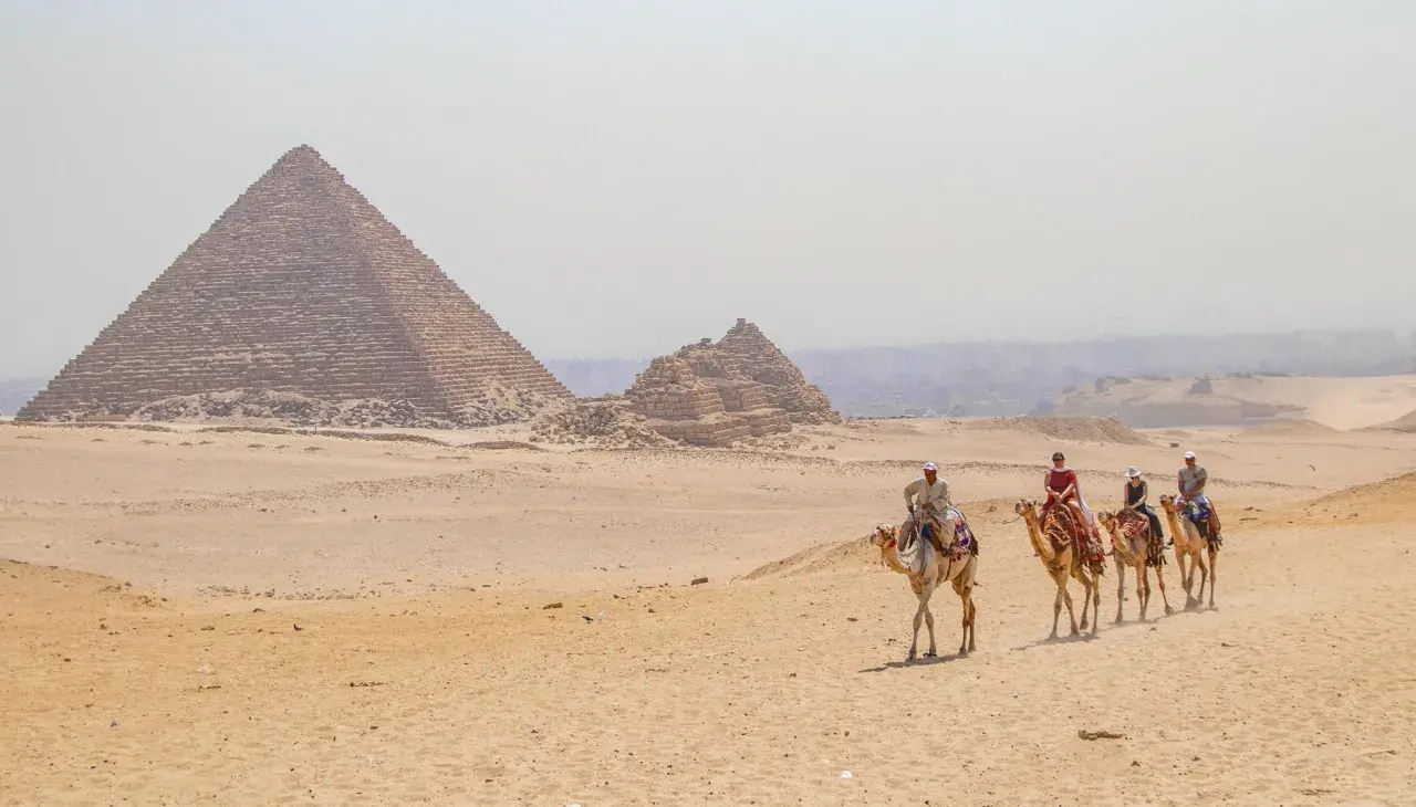 Un viaje a Egipto inolvidable: personas montan camellos frente a las pirámides de Giza bajo un cielo brumoso.