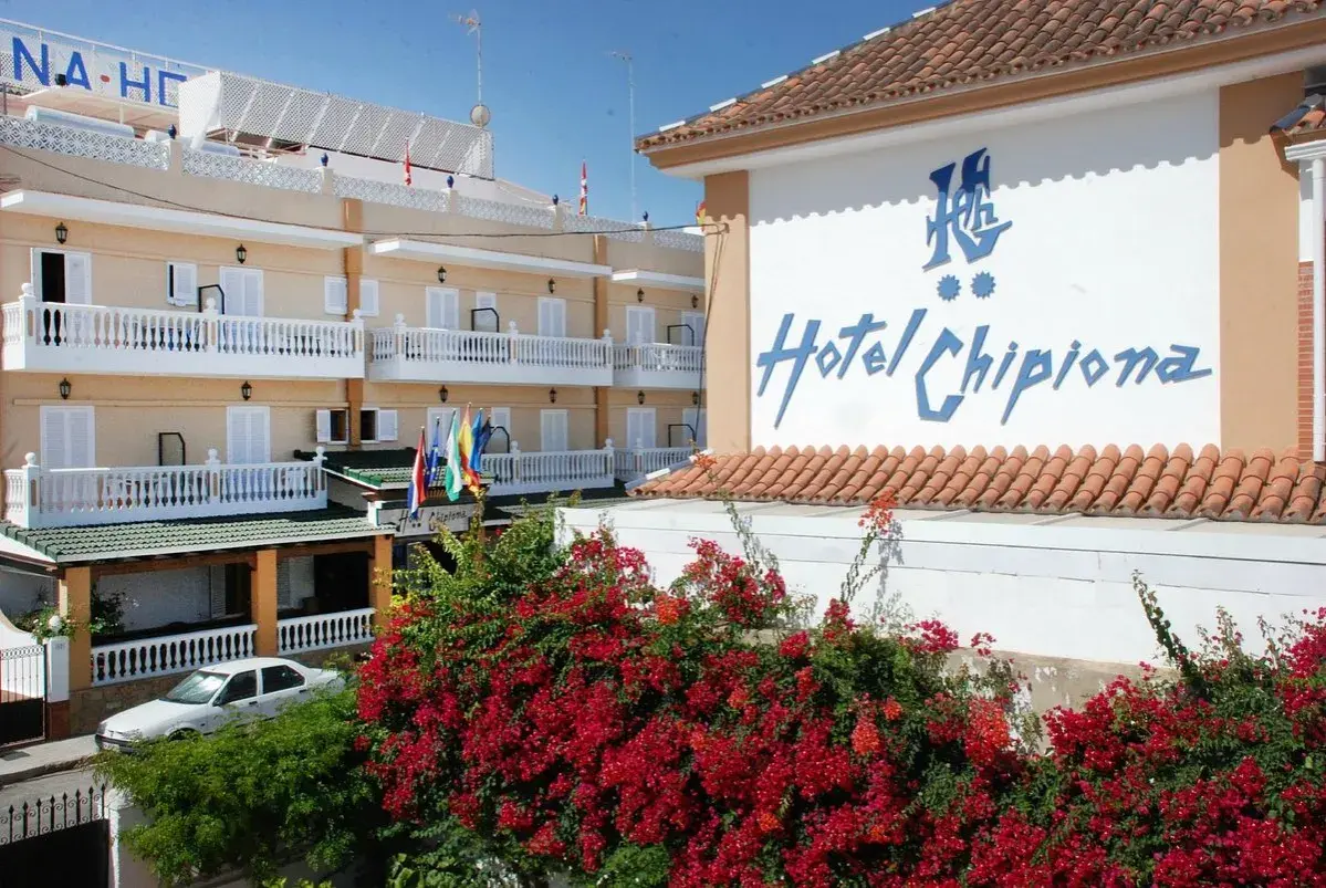 Fachada del Hotel Chipiona con balcones blancos, flores rojas y un coche aparcado.