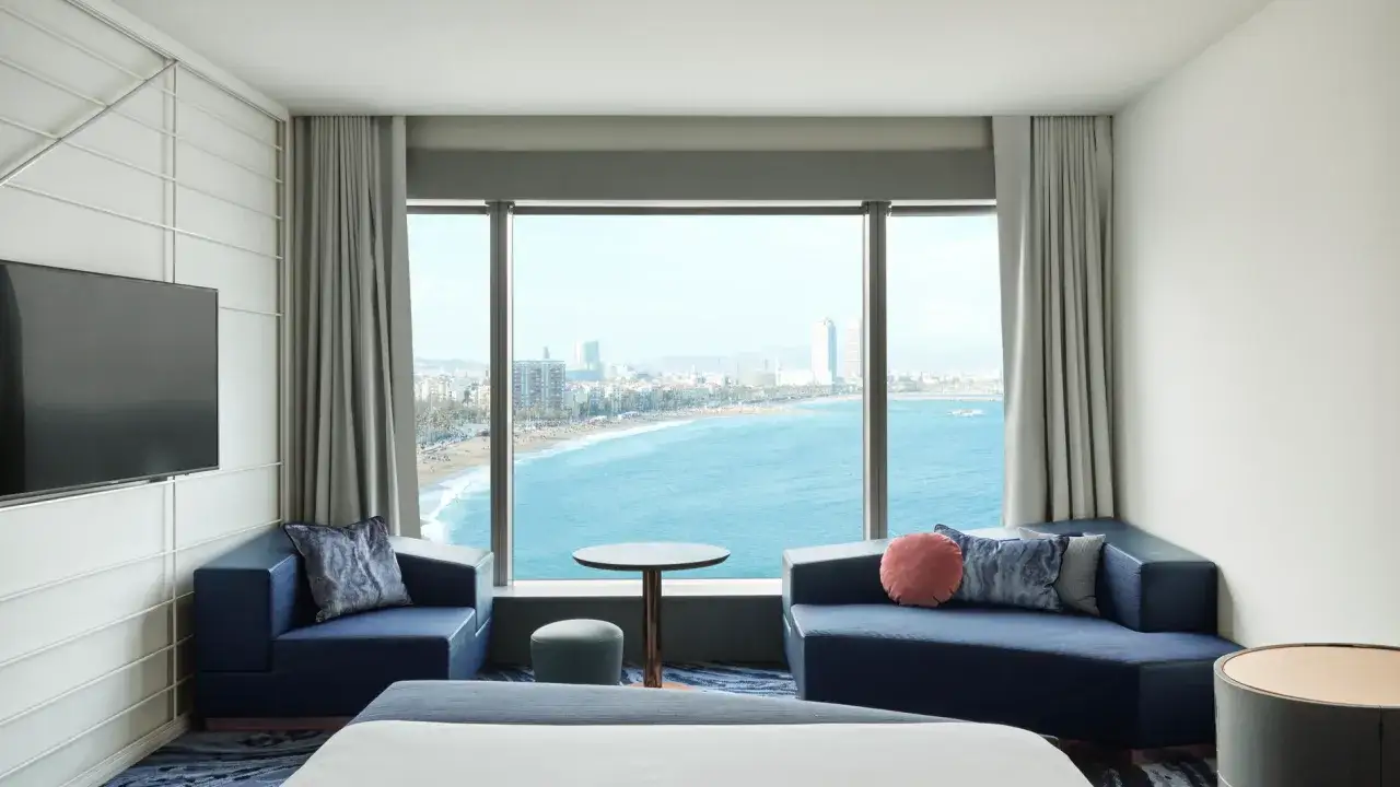 Habitación de hotel vela con vistas al mar y la ciudad. Sofá azul, sillón, mesa y televisión.