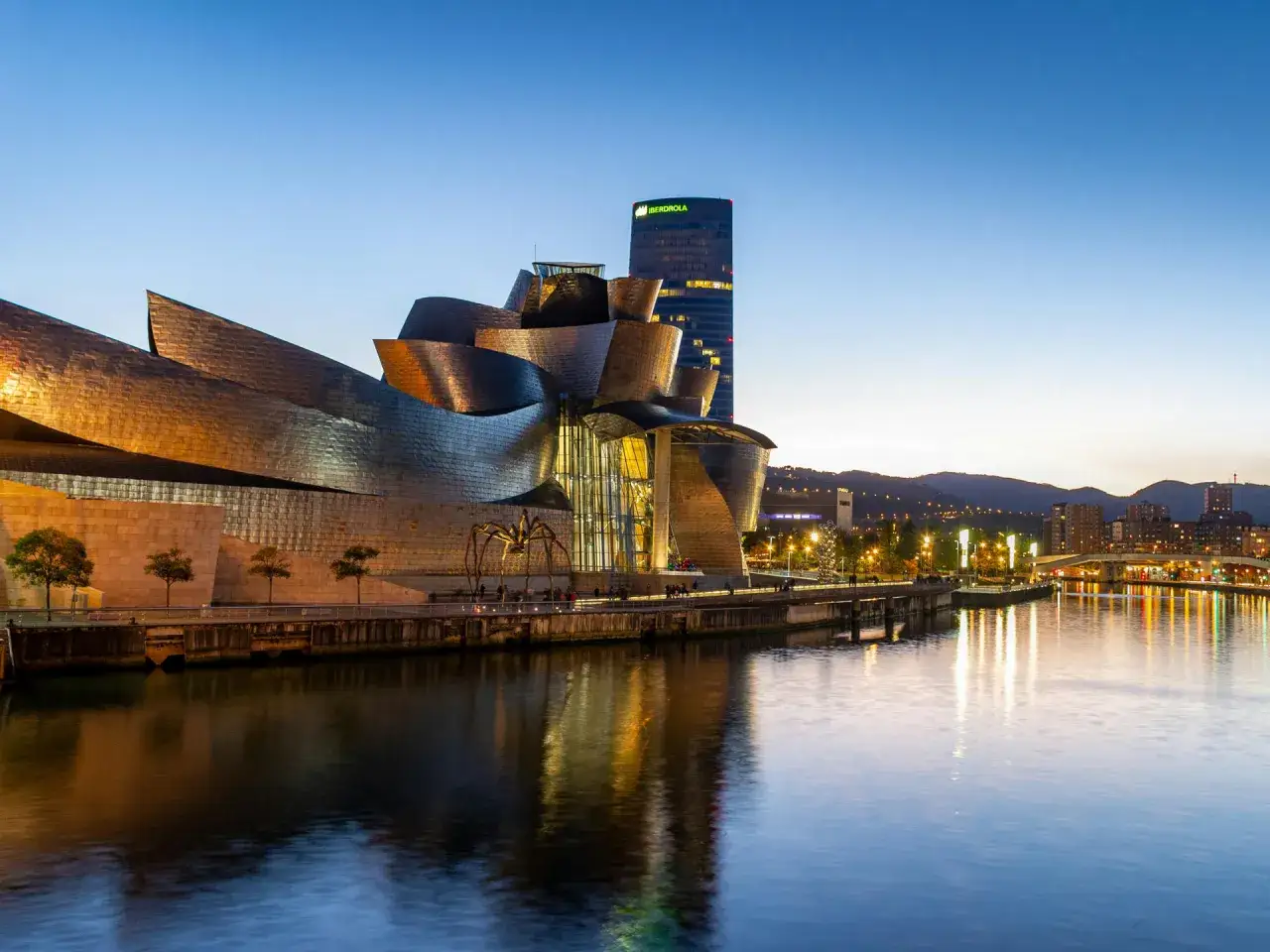 El Museo Guggenheim de Bilbao, con su icónica araña Maman, se ilumina al atardecer. Cerca, el hotel Ilunion Bilbao se alza moderno.