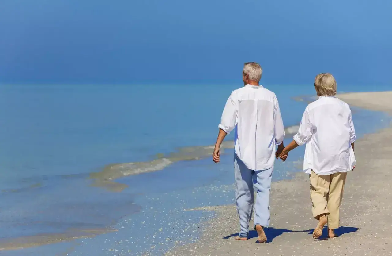 Pareja mayor pasea de la mano por la playa, disfrutando de unas vacaciones perfectas. Viajes El Corte Inglés mayores 60 años.