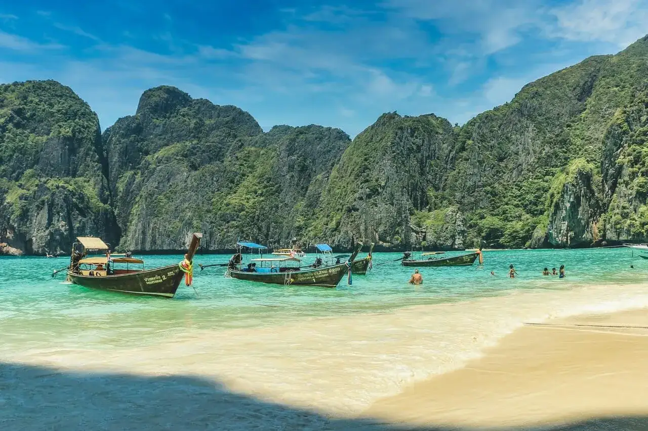 Un viaje a Tailandia: barcos tradicionales flotan en aguas turquesas frente a acantilados verdes.