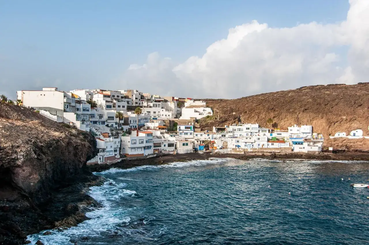 Pueblo blanco de casas apiñadas en la costa rocosa de Tufia, con el mar azul y olas rompiendo.