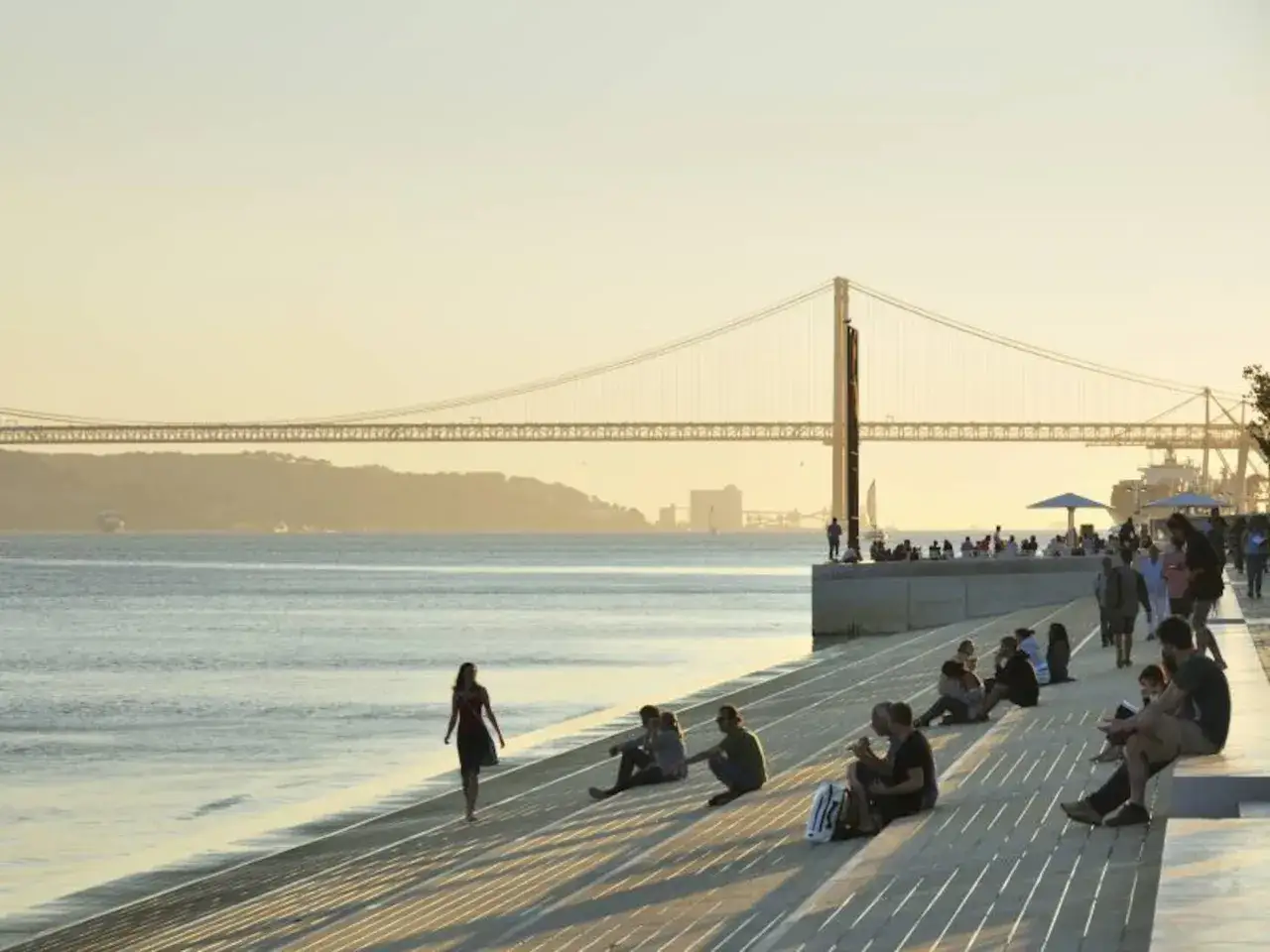 Gente disfrutando del atardecer en la ribera de Lisboa, con el puente 25 de Abril de fondo. Un lugar ideal que ver en Lisboa.