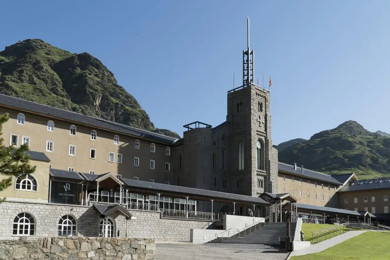 Edificio histórico en Queralbs, con torre de piedra y montañas verdes de fondo.