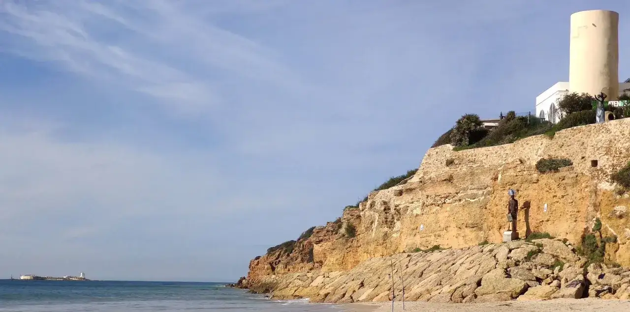 Acantilado rocoso con esculturas y un faro en la cima, bajo un cielo azul. El mar Mediterráneo baña la costa, con una estructura flotante a lo lejos. Sol Sancti Petri.