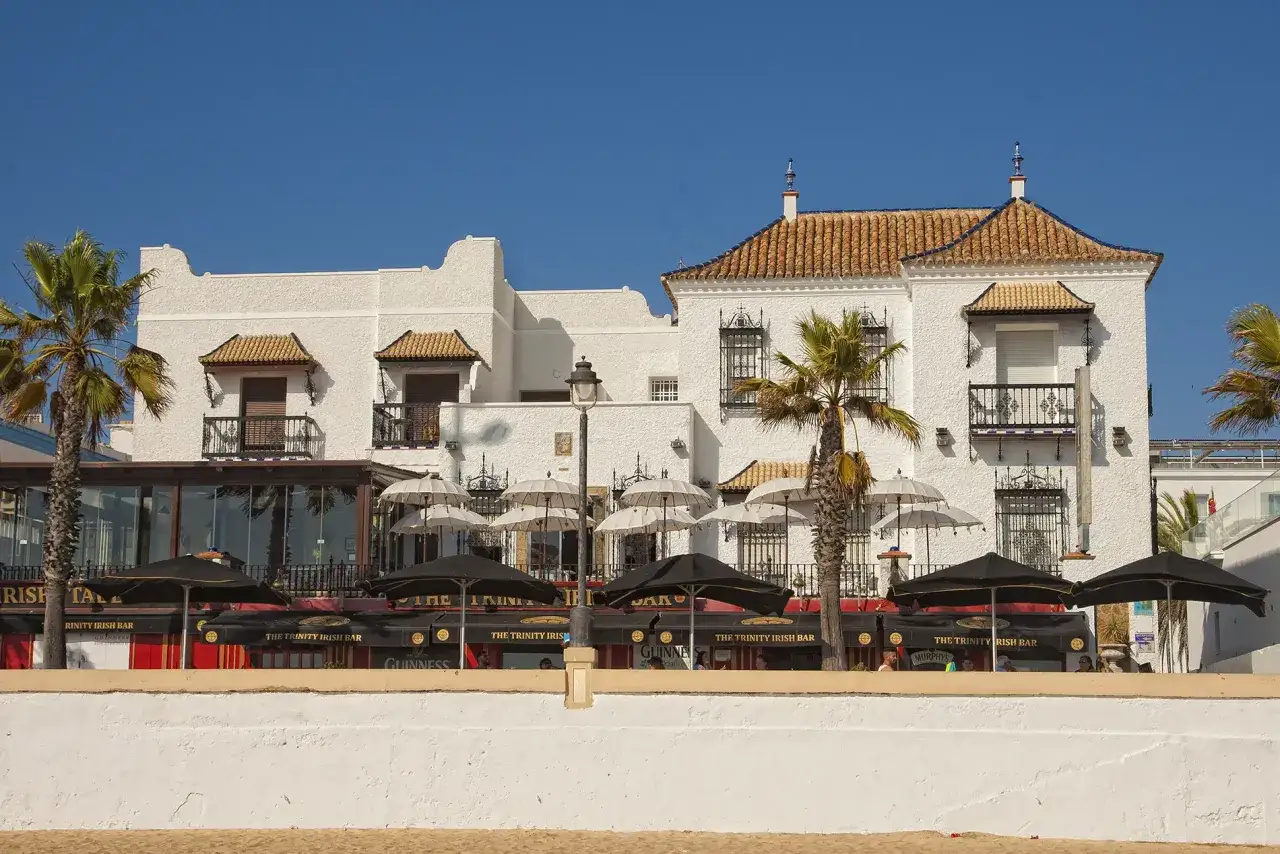 Edificio blanco con tejas naranjas, palmeras y un bar irlandés en Chipiona. Un hotel perfecto para disfrutar del sol.