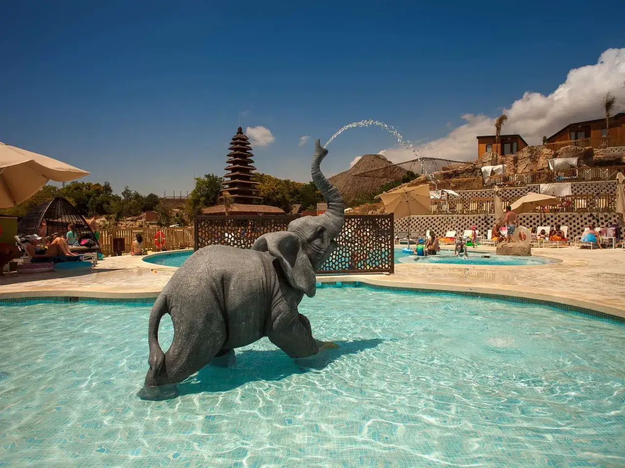 Elefante de fuente en piscina de Magic Natura Resort. Chalets de madera y gente relajándose bajo el sol.