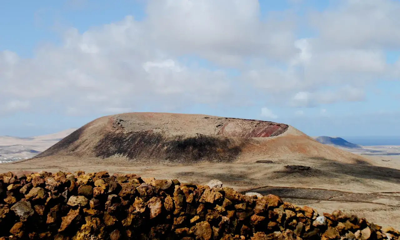 Muro de piedra volcánica en primer plano, con un cráter en la ladera de una montaña al fondo, paisaje de lajares Fuerteventura.